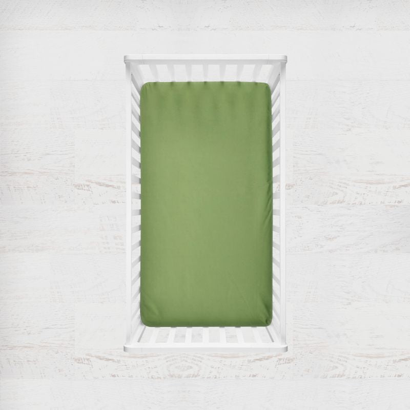 An grass green fitted crib sheet displayed on a white wooden crib, set against a light, textured white wall background. Grass Green Crib Sheet - The Creative Raccoon