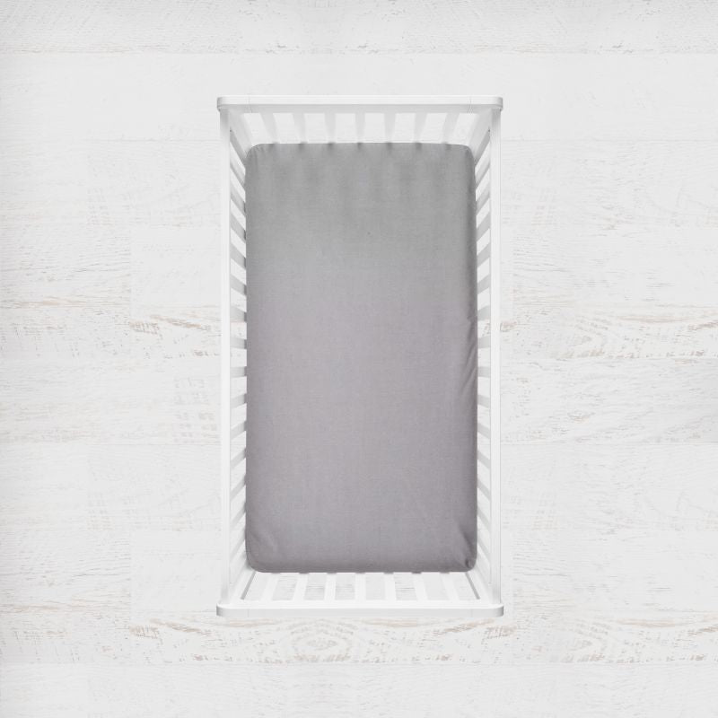 A gray fitted crib sheet shown from above inside a white wooden crib, set against a light textured white wall background. Gray Crib Sheet - The Creative Raccoon