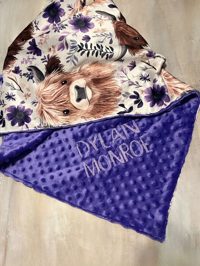 Blanket with highland cow design and floral patterns, featuring the name 'Dylain Monroe' on a purple background. Highland Cow Baby Blanket - The Creative Raccoon