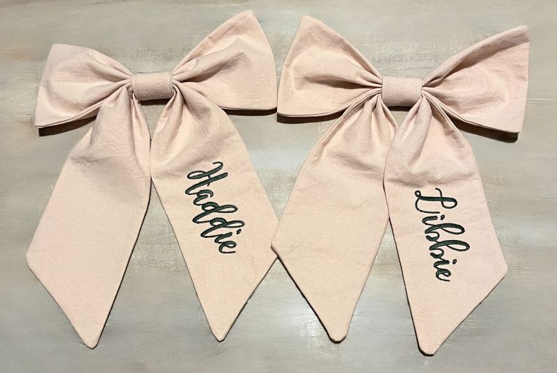 Two blush pink fabric bows with long tails, each embroidered baby's name. 
 Large Personalized Bow, Accessory - The Creative Raccoon