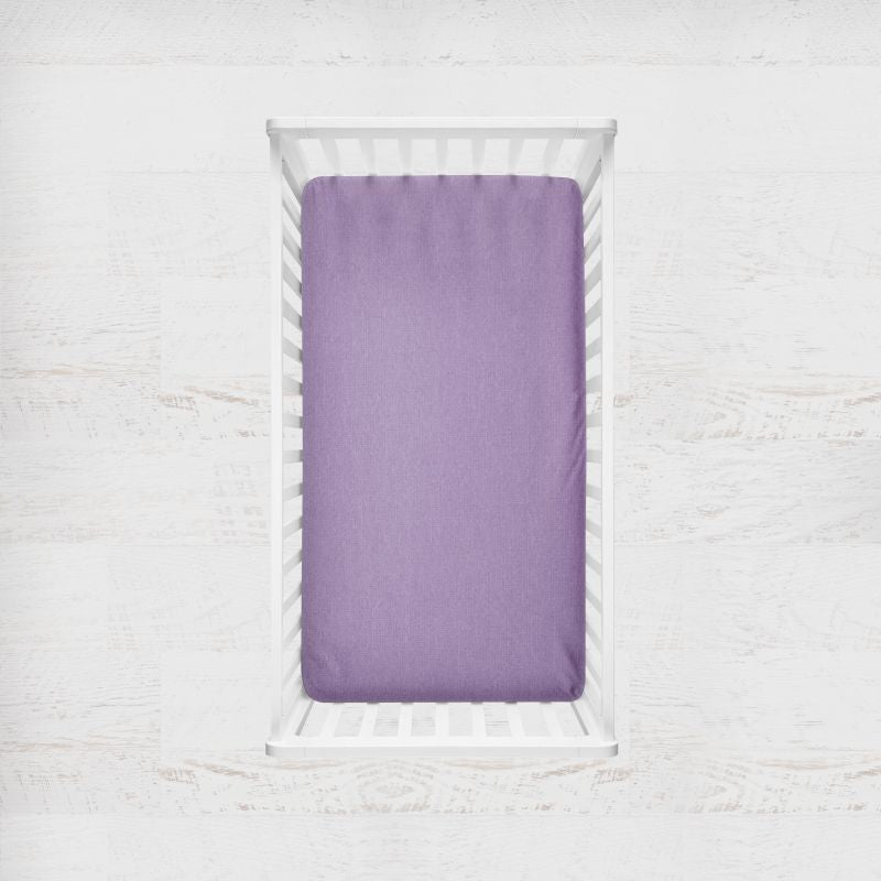 A lavender purple fitted sheet on a mattress inside a white wooden crib, photographed from a top-down view against a white textured background. Iris Purple Crib Sheet - The Creative Raccoon