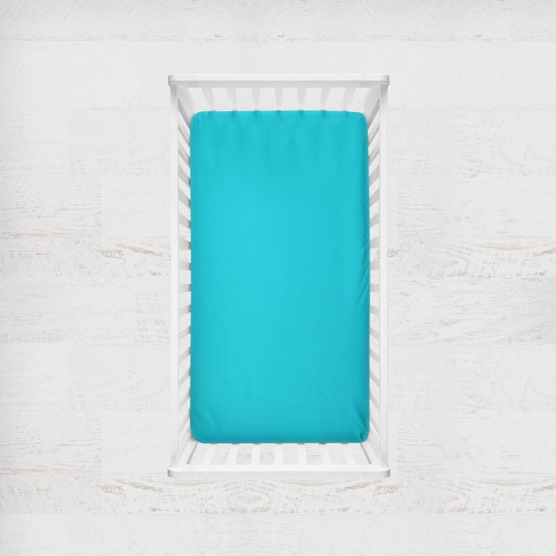 A bright turquoise fitted sheet on a mattress inside a white wooden crib, shown from a top-down view against a white textured wall. Lagoon Teal Blue Crib Sheet - The Creative Raccoon