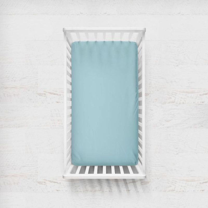 A light aqua blue fitted crib sheet displayed on a mattress inside a white wooden baby crib, set against a white textured wooden wall background. Light Aqua Blue Crib Sheet - The Creative Raccoon