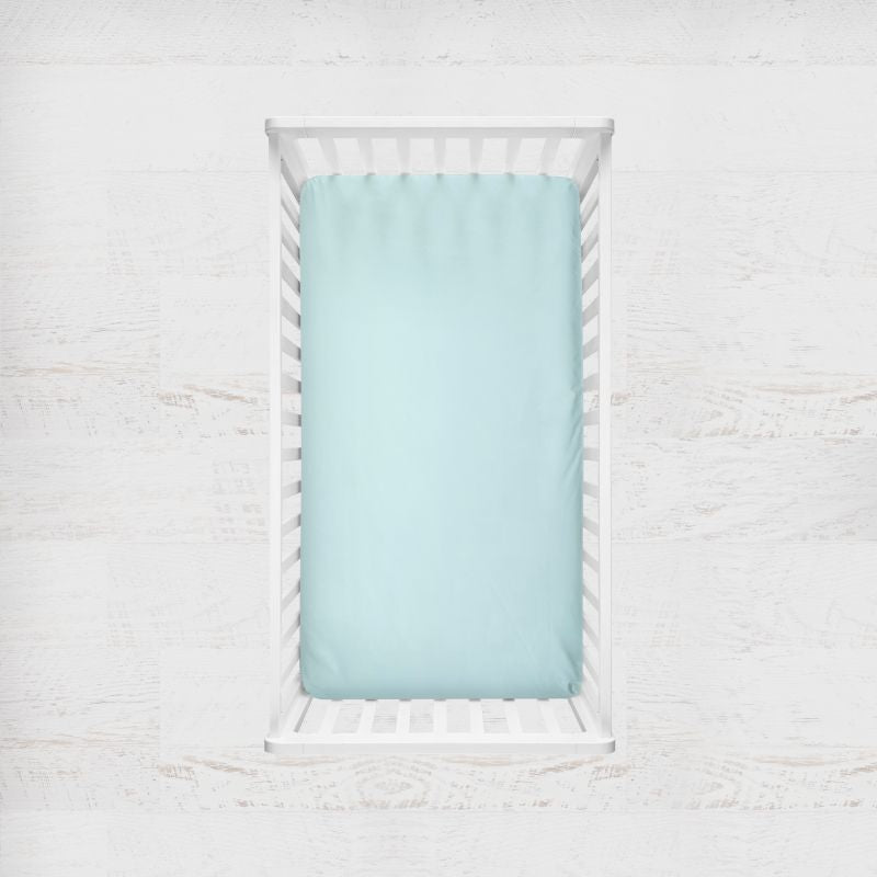 A light aqua blue crib fitted crib sheet shown inside a white wooden baby crib, set against a white textured wall background. Light Aqua Crib Sheet - The Creative Raccoon 