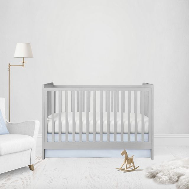 A light gray wooden baby crib with vertical slats, styled in a bright, minimalist nursery with a white armchair, gold floor lamp, and a small wooden rocking horse. Crib Bed Skirt shown in flat or straight option. 
Light Baby Blue Crib Skirt - The Creative Raccoon 