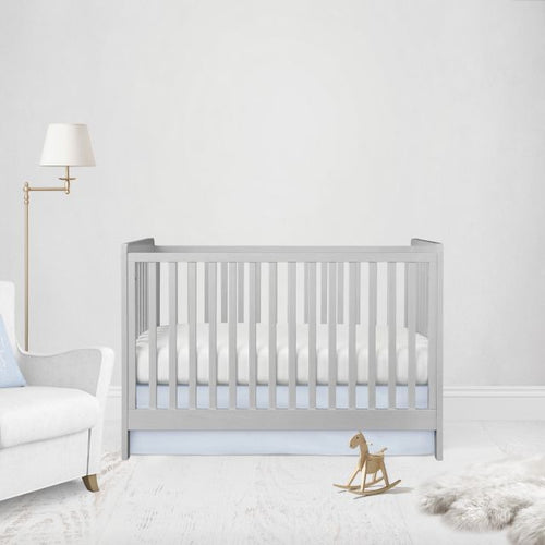 A light gray wooden baby crib with vertical slats, styled in a bright, minimalist nursery with a white armchair, gold floor lamp, and a small wooden rocking horse. Crib Bed Skirt shown in flat or straight option. 
Light Baby Blue Crib Skirt - The Creative Raccoon 