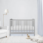 A light gray wooden crib with white bedding in a bright, modern nursery setting with a white armchair, floor lamp, and a small wooden rocking horse. 
Light Baby Blue Pleat Crib Skirt - The Creative Raccoon 