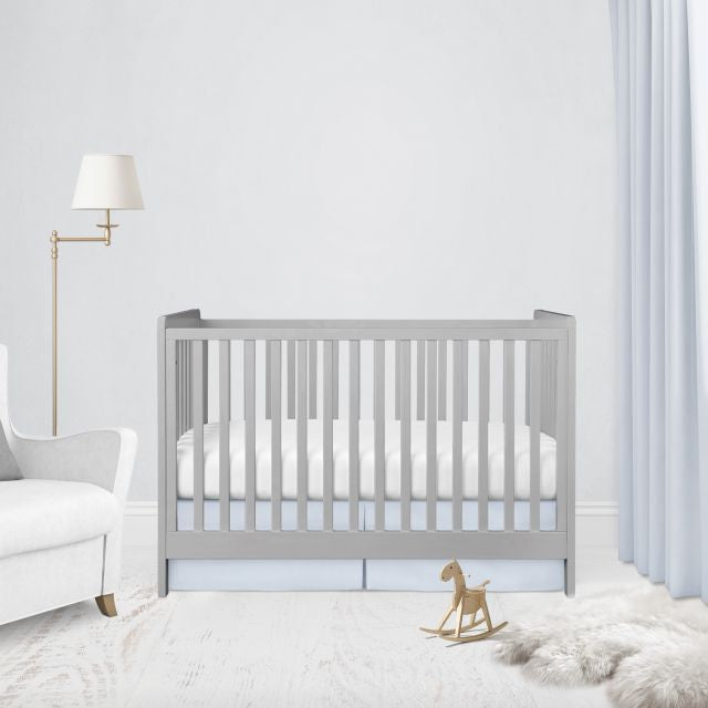 A light gray wooden crib with white bedding in a bright, modern nursery setting with a white armchair, floor lamp, and a small wooden rocking horse. 
Light Baby Blue Pleat Crib Skirt - The Creative Raccoon 