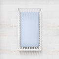 A light blue fitted sheet on a crib mattress inside a white wooden baby crib, shown from a top-down view against a white textured wall. Light Blue Crib Sheet - The Creative Raccoon