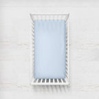 A light blue fitted sheet on a crib mattress inside a white wooden baby crib, shown from a top-down view against a white textured wall. Light Blue Crib Sheet - The Creative Raccoon