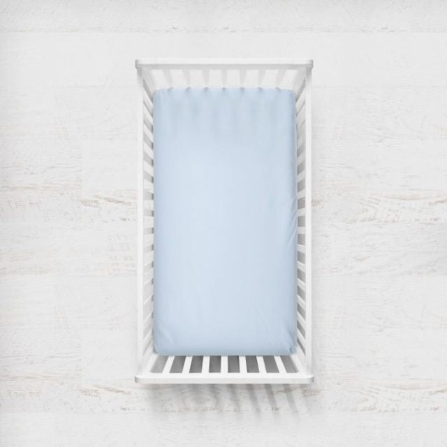 A light blue fitted sheet on a crib mattress inside a white wooden baby crib, shown from a top-down view against a white textured wall. Light Blue Crib Sheet - The Creative Raccoon