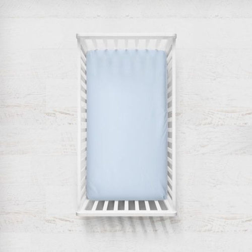 A light blue fitted sheet on a crib mattress inside a white wooden baby crib, shown from a top-down view against a white textured wall. Light Blue Crib Sheet - The Creative Raccoon