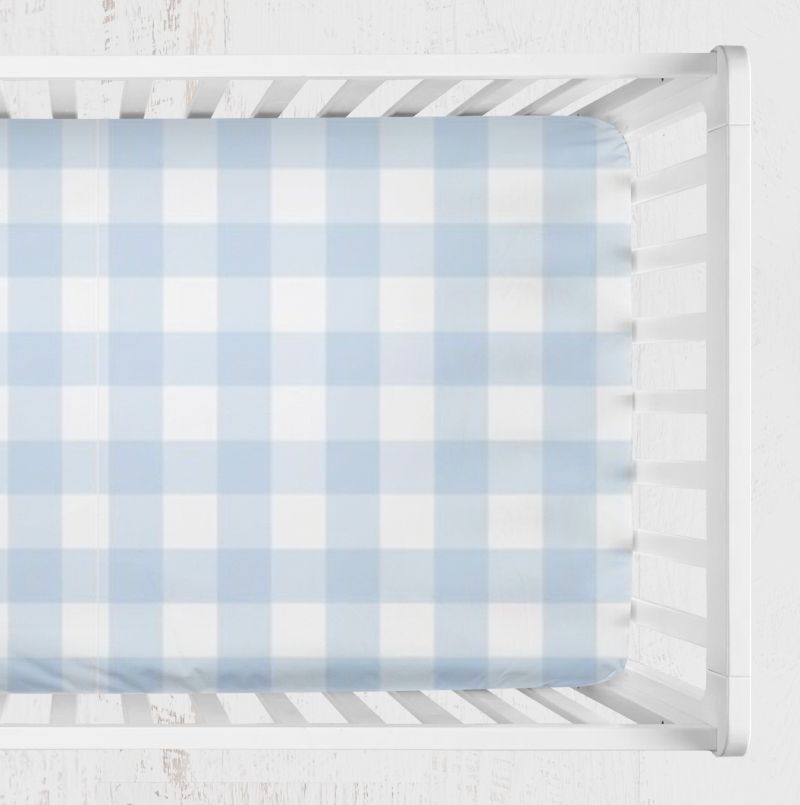A light blue and white gingham crib sheet fitted inside a white wooden crib, shown from a top-down view against a white textured background. Light Blue Gingham Crib Sheet - The Creative Raccoon