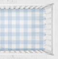 A light blue and white gingham crib sheet fitted inside a white wooden crib, shown from a top-down view against a white textured background. Light Blue Gingham Crib Sheet - The Creative Raccoon