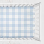 A light blue and white gingham crib sheet fitted inside a white wooden crib, shown from a top-down view against a white textured background. Light Blue Gingham Crib Sheet - The Creative Raccoon