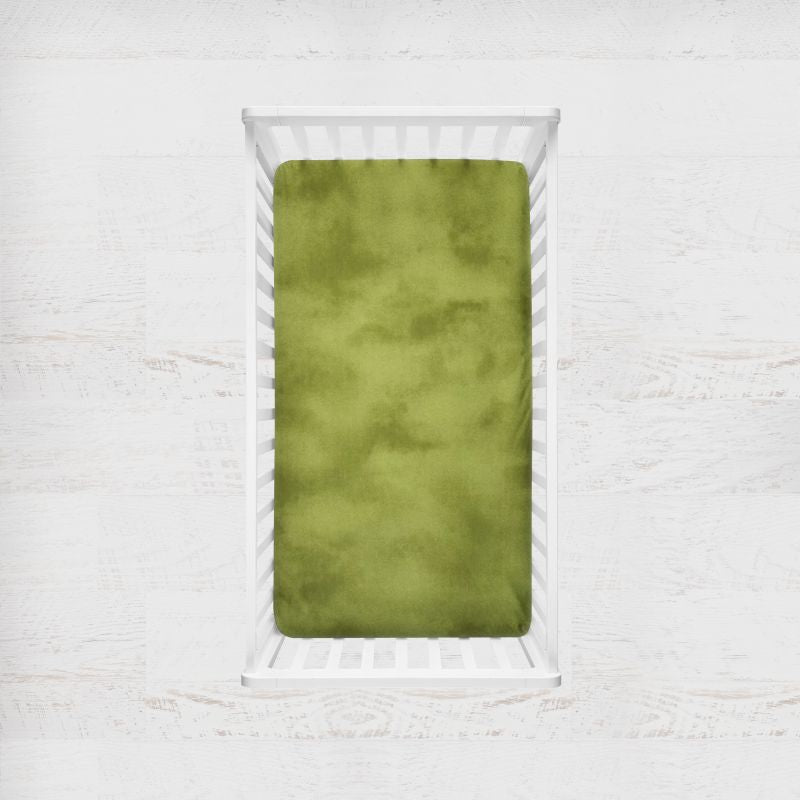 An light olive-green textured crib sheet displayed inside a white wooden baby crib, set against a light, rustic white wood wall background. Light Olive Green Crib Sheet - The Creative Raccoon