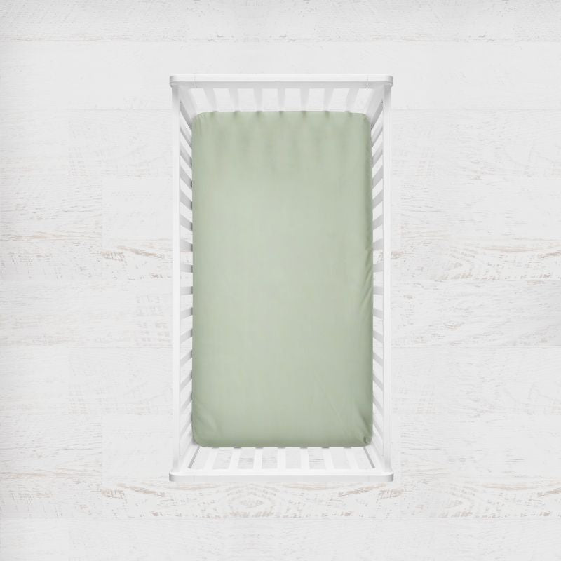 A light sage green fitted crib sheet displayed inside a white wooden crib, set against a light, textured white wall background. Light Sage Green Crib Sheet - The Creative Raccoon
