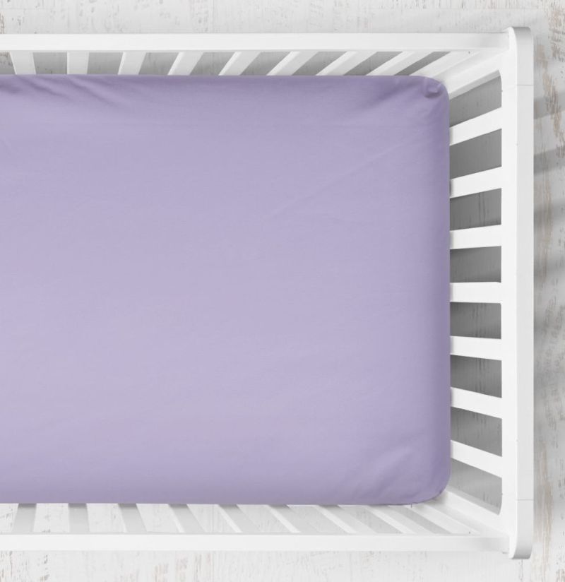 A lavender purple fitted crib sheet on a mattress inside a white wooden crib, shown from a top-down perspective. Lilac Crib Sheet - The Creative Raccoon