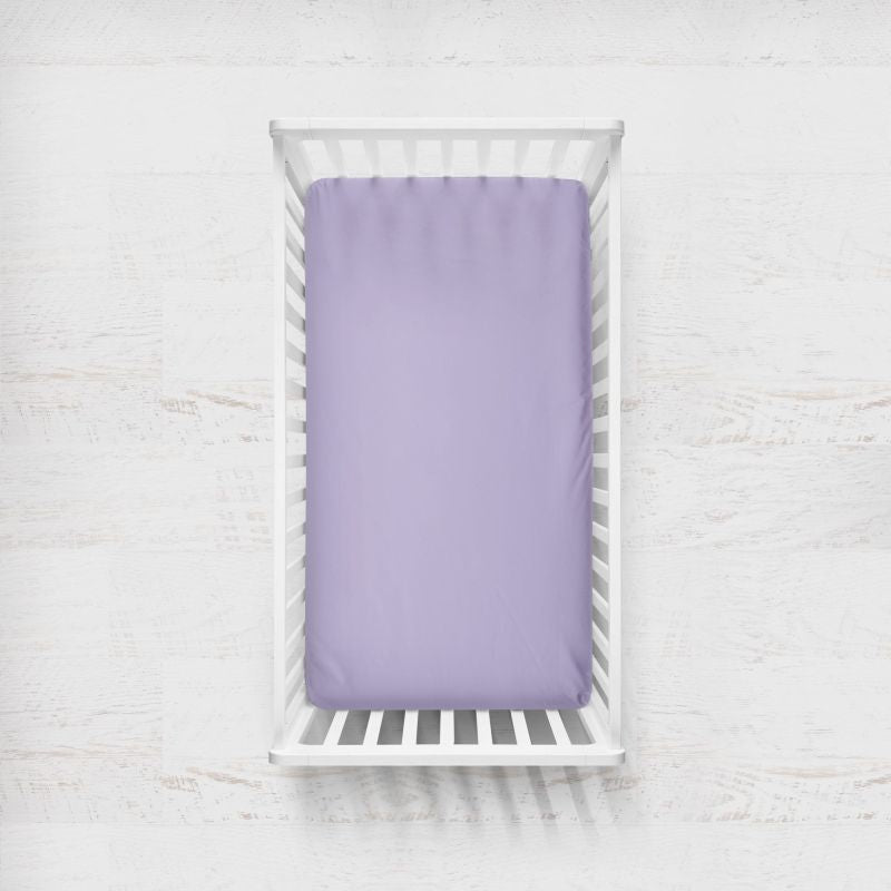 A lilac purple fitted sheet on a mattress inside a white wooden baby crib, photographed from a top-down perspective on a light, textured white background. Lilac Light Purple Crib Sheet - The Creative Raccoon