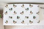 A white changing pad or mattress cover printed with a repeating pattern of mallards in different poses, shown resting on a beige carpeted floor. Mallard Duck Changing Pad Cover - The Creative Raccoon