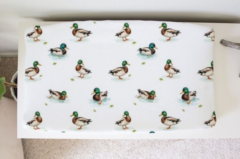 A white changing pad or mattress cover printed with a repeating pattern of mallards in different poses, shown resting on a beige carpeted floor. Mallard Duck Changing Pad Cover - The Creative Raccoon