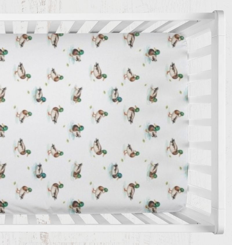 A white crib fitted sheet with a repeating pattern of mallard ducks, shown inside a white wooden crib. Mallard Duck Crib Sheet - The Creative Raccoon