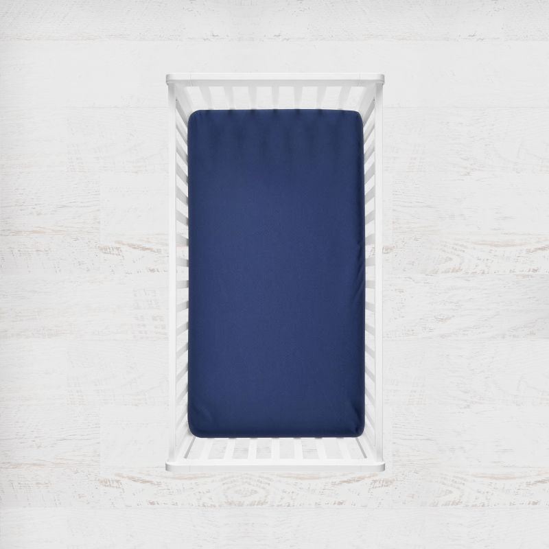A navy blue fitted crib sheet displayed on a mattress inside a white wooden crib, set against a light, textured white wall background. Marine Navy Blue Crib Sheet - The Creative Raccoon