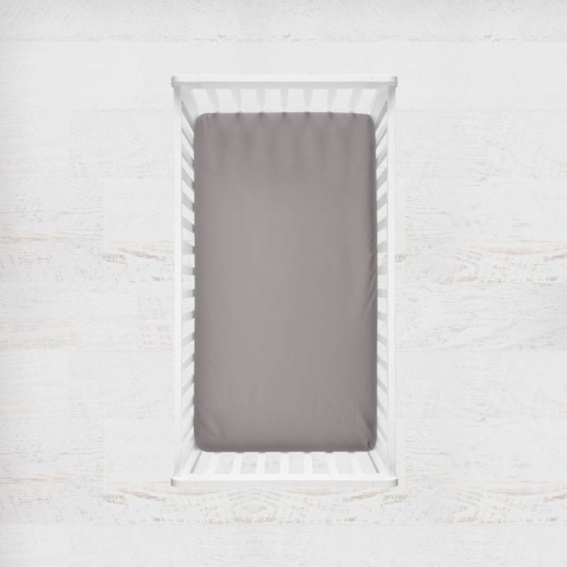 A top-down view of a white wooden crib with a fitted sheet in a medium gray color, set against a white distressed wood background. Medium Gray Crib Sheet - The Creative Raccoon