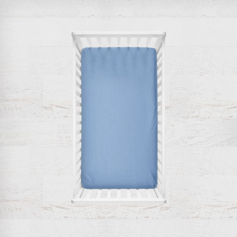 A light blue fitted crib sheet displayed inside a white wooden baby crib, shown from a top-down view against a white textured background. Monet Light Blue Crib Sheet - The Creative Raccoon