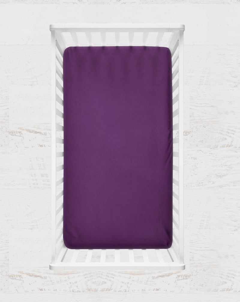 A solid purple fitted crib sheet shown from a top-down view inside a white wooden crib against a white textured wall. Mulberry Purple Crib Sheet - The Creative Raccoon