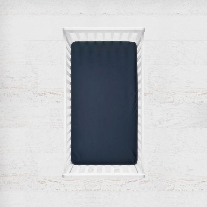 A navy blue fitted crib sheet displayed inside a white wooden baby crib, set against a light, textured white wall background. Navy Blue Crib Sheet - The Creative Raccoon 