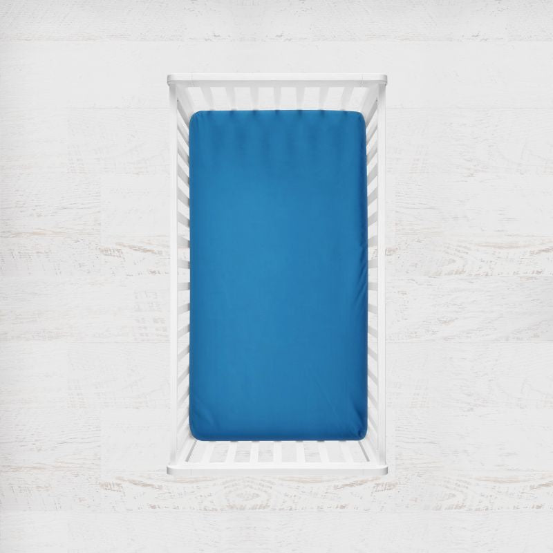 A bright blue fitted sheet on a mattress inside a white wooden crib, shown from a top-down view against a white textured wall. Pacific Bright Blue Crib Sheet - The Creative Raccoon