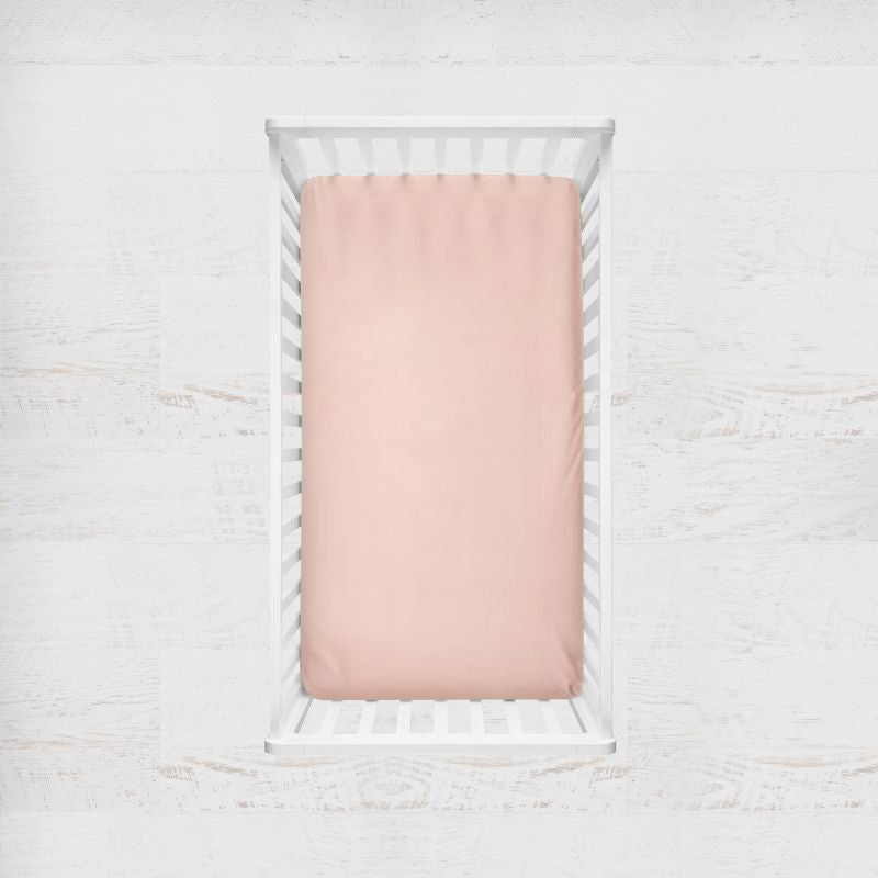 A pale blush pink fitted sheet displayed inside a white wooden baby crib, shown from a top-down view against a white textured background. Peach Crib Sheet - The Creative Raccoon