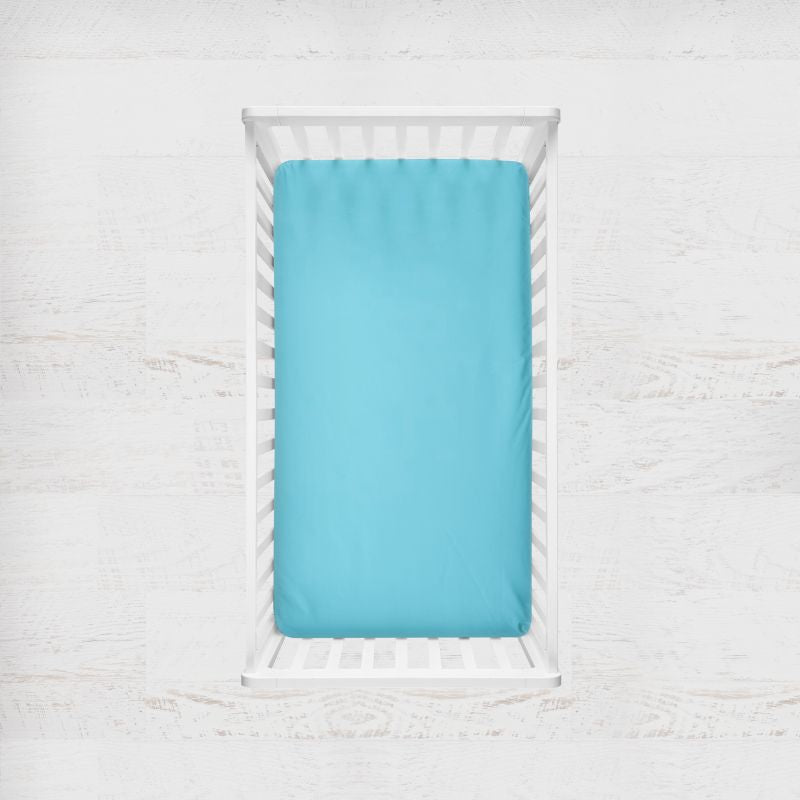 A bright turquoise blue fitted sheet on a mattress inside a white wooden crib, shown from a top-down view against a white textured background. Peacock Light Teal Blue Crib Sheet - The Creative Raccoon