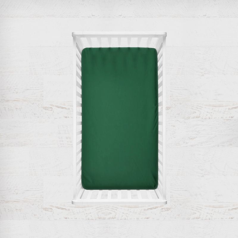 A dark green fitted sheet on a mattress inside a white wooden crib, shown from a top-down view against a white textured wall. Pesto Green Crib Sheet - The Creative Raccoon