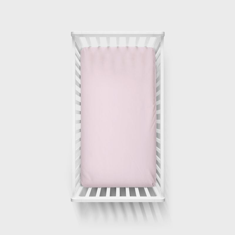 A light petal pink fitted sheet displayed on a white wooden baby crib, shown from a top-down perspective against a light gray background. Petal Pink Crib Sheet - The Creative Raccoon