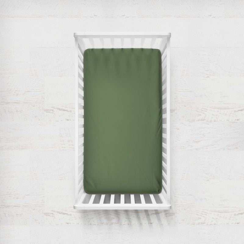 A pine sage green fitted crib sheet displayed on a mattress inside a white wooden baby crib, set against a light, textured white wall background. Pine Green Crib Sheet - The Creative Raccoon