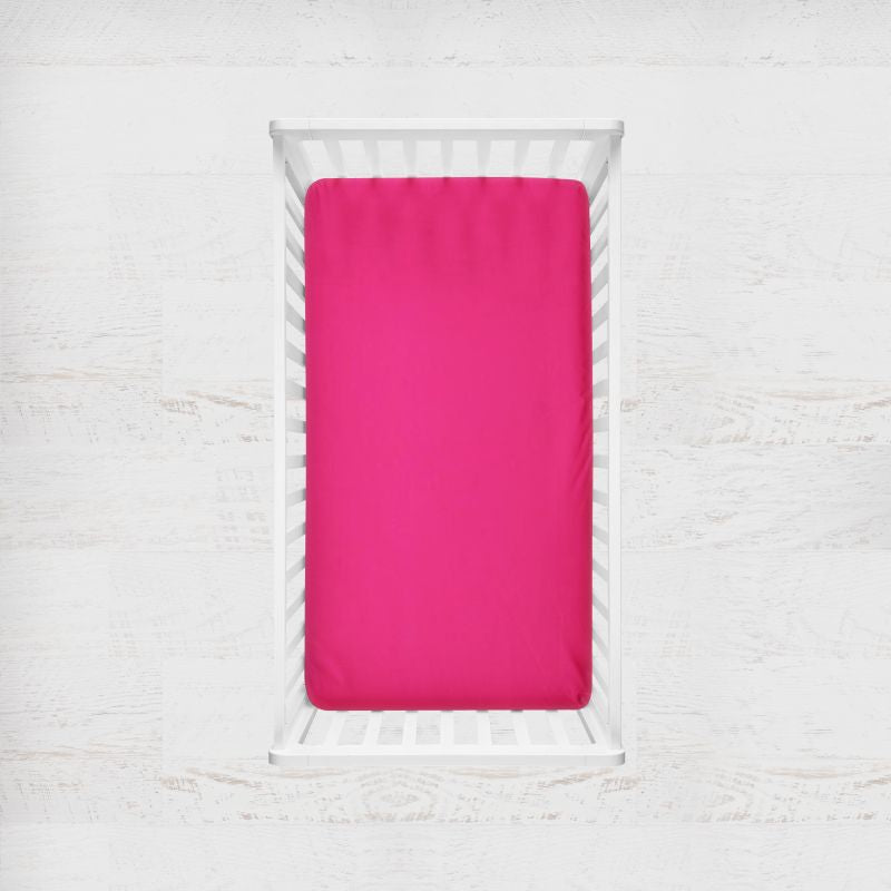 A bright pink fitted sheet on a mattress inside a white wooden crib, shown from a top-down view against a white textured wall. Pomegranate Bright Pink Crib Sheet - The Creative Raccoon