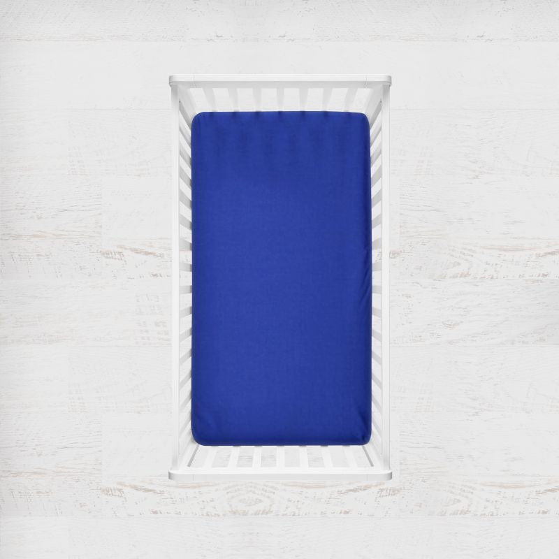 A royal blue fitted crib sheet displayed on a mattress inside a white wooden crib, set against a light, textured white wall background. Royal Blue Crib Sheet - The Creative Raccoon 