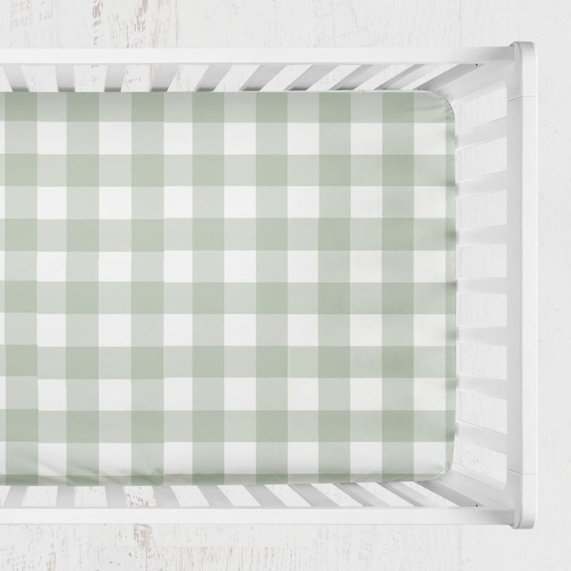 A fitted crib sheet with a sage green and white gingham check pattern, shown inside a white wooden crib. Sage Gingham Crib Sheet - The Creative Raccoon 
