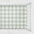A fitted crib sheet with a sage green and white gingham check pattern, shown inside a white wooden crib. Sage Gingham Crib Sheet - The Creative Raccoon 