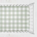 A fitted crib sheet with a sage green and white gingham check pattern, shown inside a white wooden crib. Sage Gingham Crib Sheet - The Creative Raccoon 