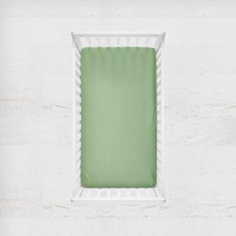 A sage green fitted crib sheet displayed inside a white wooden crib, set against a light, textured white wall background. Sage Green Crib Sheet - The Creative Raccoon