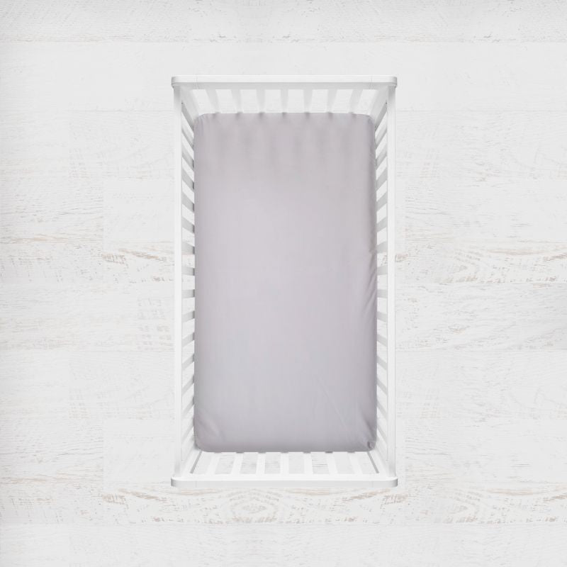 A light silver-gray crib sheet fitted inside a white wooden crib, shown from a top-down view against a white textured background. Silver Gray Crib Sheet - The Creative Raccoon