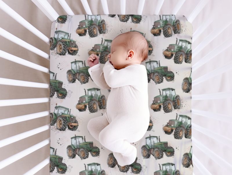 A sleeping newborn baby in a white footed onesie lies on a fitted crib mattress with a green tractor patterned cover, photographed from above inside a white crib.
Tractor Crib Sheet with Cream Background - The Creative Raccoon