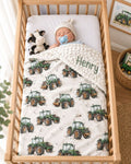 A baby sleeping in a wooden crib, wrapped in a personalized cream blanket printed with green tractor illustrations and the name 'Henry' embroidered on the corner. The baby wears a white knit hat and has a blue pacifier, with a plush cow toy beside them. Tractor Minky Blanket with Cream Background - The Creative Raccoon