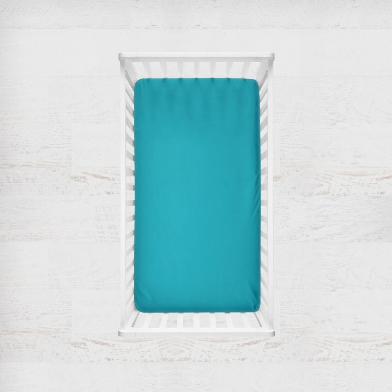 A bright turquoise fitted sheet on a mattress inside a white wooden crib, shown from a top-down view against a white textured wall. Turquoise Dark Teal Blue Crib Sheet - The Creative Raccoon