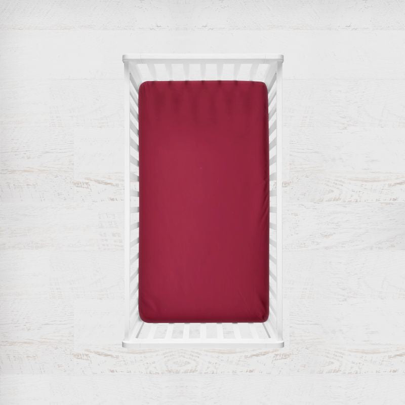 Red fitted sheet on a white bed frame against a white textured wall. ruby red crib sheet - the creative raccoon