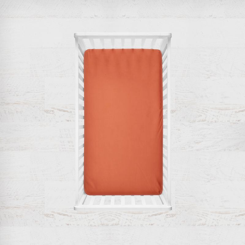 Orange fitted sheet on a white crib with a light gray background. terracotta orange crib sheet - the creative raccoon