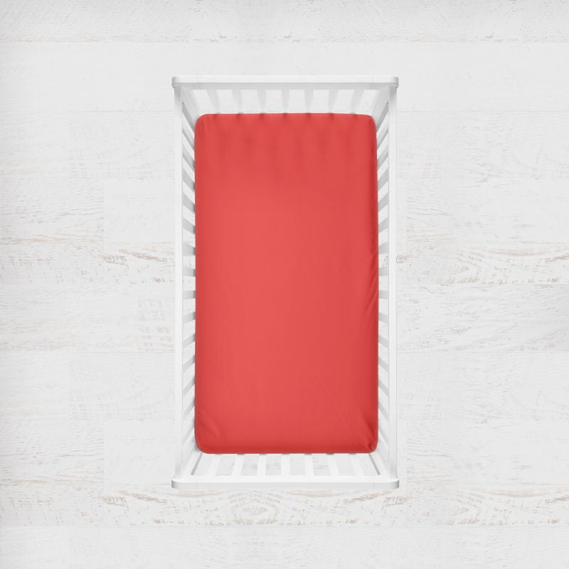 Red fitted sheet on a white bed with a light gray background. Tomato red crib sheet - the creative raccoon